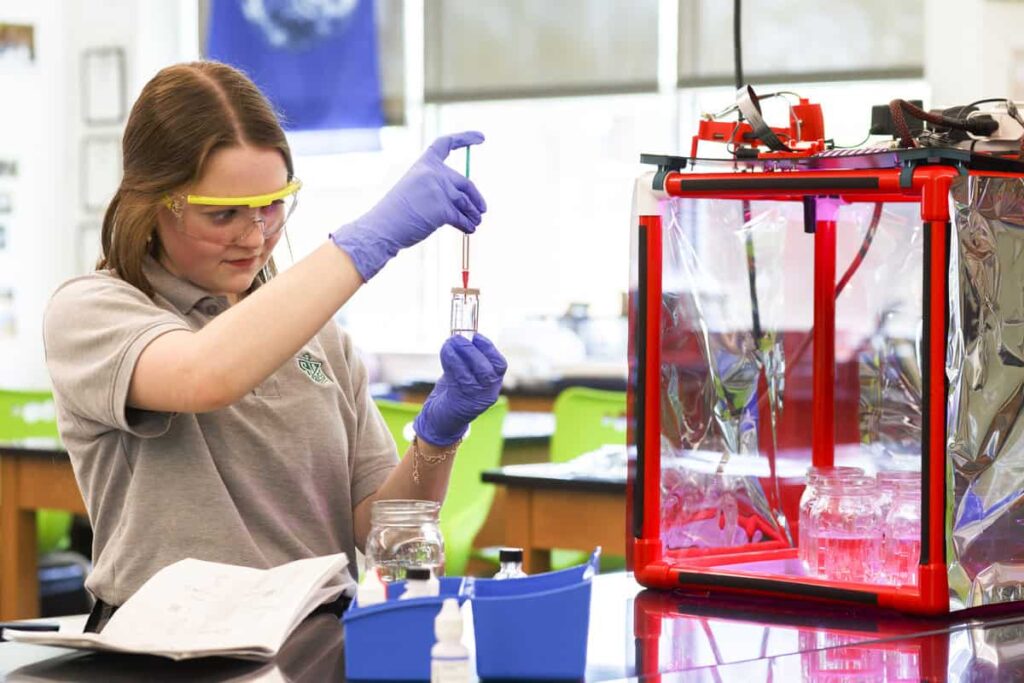 A girl wearing safety glasses inserts a pipette into a glass container.