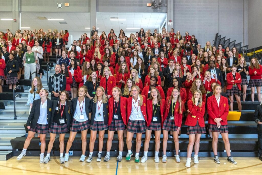 Girls in school uniforms link arms in the bleachers during an assembly.