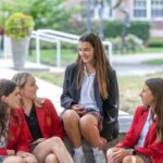 Four Academy students wearing school uniform blazers chat outside on a sunny day.