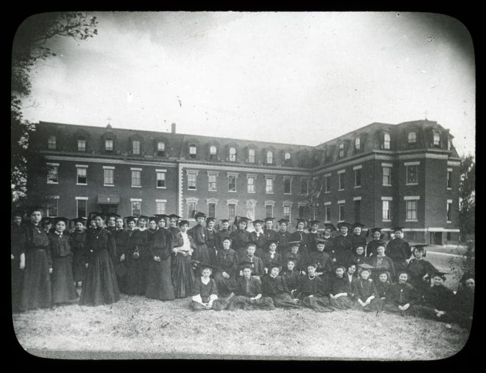 Black and white photo of graduates with cap and gown in front of a college building.